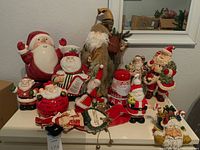 Full overview of assorted Santa figurines including cookie jars, soap dispenser, plush figure, and teapot on white table in front of mirror.
