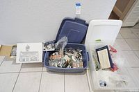 Overview of two plastic storage bins with various Department 56 Christmas Snow Village items and Delft blue ornament inside, along with boxes beside them.