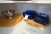 Three cobalt blue rectangular glass bakeware dishes stacked on one of two round wooden Lazy Susan turntables; metal perforated strainer with wooden base on other turntable.