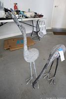 Pair of metal crane garden sculptures on a garage floor, one crane with neck raised and the other with neck lowered.