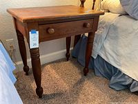 Dark walnut nightstand showing front with one drawer and turned legs, positioned beside a bed.
