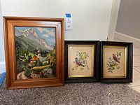 Three pieces of wall art: a mountain scene cross stitch piece with a natural wooden frame and two matching framed bird prints by Vincent with black frames and gold inner detail.