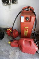 Five gasoline cans of various sizes arranged on concrete floor in garage. One large can has a hose attached, others have yellow caps; all red with signs of use.