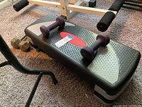 Fitness step platform with two purple 5 lb dumbbells resting on top and 12 lb metal dumbbells on the floor.