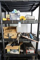 Photo of garage shelves holding all items including boxed floor jack, tools in cardboard boxes, battery charger, and cloths