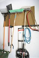 Photo of assorted gardening tools hanging on pegboard including rakes, shovels, brooms, a garden hose, a bow saw, and portable dolly.
