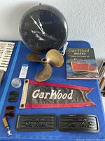 Overview of all GarWood boat related items on blue surface, including clock, propeller, book, banner, plates, glass cover, vents, and spark plug.