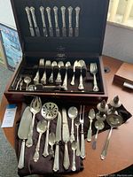Full view of open wood jeweler’s box with eight knives in lid and assorted flatware and serving pieces in base
