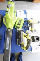 Top-down view of the Greenworks leaf blower, Ryobi drill, circular saw, chargers, and instruction manuals on a blue table.