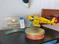 Yellow Stearman U-51 model biplane with black display stand and postcard in clear holder next to a round collectible tin