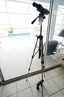 Velbon monocular mounted on tripod, standing near sliding glass door with pool visible outside. Included binoculars resting on floor beside tripod.