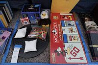 Full layout of Monopoly Mickey Mouse 75th Anniversary board game box with its lid showing the game title and artwork. Includes a plush eagle toy, a bag of varied colorful plastic pieces, two small white cushions, a black circular item, and a Bicycle playing card deck all positioned around the game box.