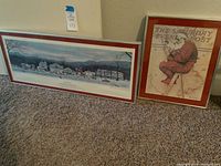 Both framed prints set on carpet floor, one large panoramic winter village, one portrait Santa Claus image