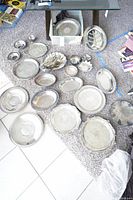 Overview of varied silver plated platters, plates, bowls and flatware spread on carpet and tile floor under glass table, showing different sizes and styles including oval and round shapes
