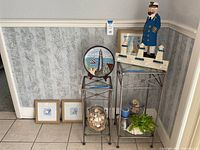 Two metal and glass side tables with various coastal theme decorations arranged including lighthouse statues, shell jars, framed artworks, and a wooden sailor figure.