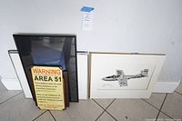 Several framed items leaning against wall on floor. Includes framed signed print of military aircraft and others.