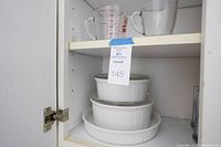 Three white CorningWare ceramic dishes stacked inside a cabinet shelf. Two casserole dishes with lids and one quiche dish on the bottom.