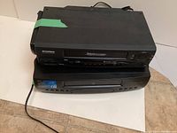Two VHS players stacked, Sylvania on top and GE below, showing front and top views with power cords and remote controls.