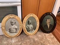 Three large oval antique picture frames displayed on a tiled floor, two cream-colored ornate frames and one dark wood frame containing vintage portraits.