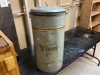Image showing the antique cylindrical flour tin standing on a table in an indoor setting, displaying its color, shape, lid, and decorative labeling.