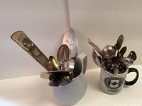 Photo showing two containers holding assorted silver serving pieces and small spoons. Left container is a white ceramic pitcher and right container is a white ceramic mug with black handle.