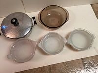 Top-down view showing one metal casserole pot with a lid and four clear glass bowls, three with lids and one without, all arranged on floor tiles.