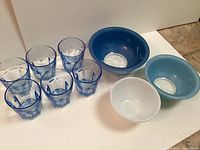 Photo showing seven faceted clear drinking glasses and three solid-colored glass bowls in blue and white shades on a white background.
