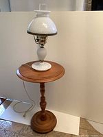 Photo showing the white hobnail glass hurricane lamp with a clear glass chimney on top of a round wooden side table with a turned central pedestal base and circular foot.