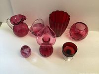 Photo of six rose-colored glass items on white background: pitcher, large vase, small vase, votive piece, basket-shaped vase, and heart-shaped decorative piece.