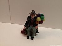Porcelain figurine of a seated elderly man holding colorful balloons and a bag beside him, on a neutral background