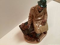 Photo of Royal Doulton Lunch Time porcelain figurine showing a man in a trench coat, black hat, sitting on a bench with several squirrels around him, holding a lantern.