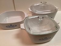 Photo showing 3 white Corning Ware casserole dishes with blue cornflower design. Two have glass lids, one is without a lid. Dishes have square shapes and integrated handles.