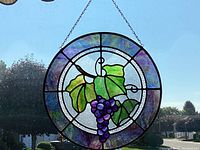 Photo showing round stained glass ornament with purple grapes and green leaves hanging in a window