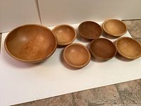 Photo of one large wooden bowl and six smaller wooden bowls arranged in front of a white backdrop on tile floor.