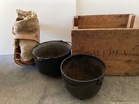 Full view of all items: wooden box, two black cast iron kettles, wicker basket with rolled burlap sack