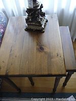 Two wooden nesting end tables, one smaller table nesting under the larger table, shown with a decorative lamp on top. Tables are medium brown with natural wood grain and knots visible.