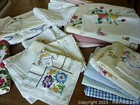 Assorted folded vintage linens displayed on a table. Visible are several hand-embroidered pieces featuring floral designs, some with lace edging.