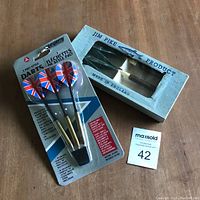 Photo showing two sets of darts: one unopened set with Union Jack flights in plastic packaging and one Jim Pike set in original box with window showing three darts with feather flights.