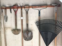 Full view of assorted gardening tools including hand trowel, cultivator, hoes, shovels, metal rake, and large plastic Anvil rake hung on wall