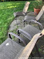 Photo showing four brown mesh and metal backyard patio lawn chairs arranged on a stone patio with grass background. Chairs have armrests and a slim profile.