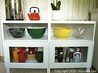 Front view of white storage cabinet with decorative items on top and inside shelves showing colorful bowls, pitchers, and glassware. The cabinet stands against wallpapered wall.