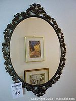 Full front view of the oval mirror showing the reflective surface and detailed metal frame with floral and leaf pattern, mounted on a wall with paintings reflected in it.