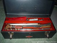 Open metal toolbox showing assorted hand tools inside, including wooden-handled screwdriver, wrench, hammer, and other tools.