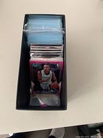 Top-down photo inside black box showing a large stack of clear plastic sleeves and assorted basketball trading cards in front.