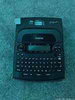 Front view of the Brother P-Touch label maker showing keyboard, screen, and installed white tape cartridge.