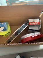 Photo showing open cardboard box with various packing supply items including yellow tape roll, adhesive label rolls, ULINE boxes, and red tape dispenser.