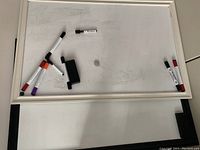 Two whiteboards laid flat with white frames, visible usage marks and smudges on the surfaces. Several dry erase markers and one black eraser placed on top of the boards.
