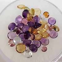 Clear photo showing various loose gemstones in shades of yellow, red, purple, and pink on a white background.