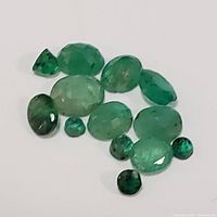 Top view of 12 loose faceted emerald gemstones in varying sizes and shapes arranged on white background.