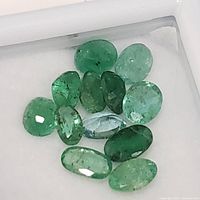 Top close-up view of eleven loose oval-cut emerald gemstones of various green shades, showing inclusions and faceting details.